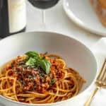 A plate of lentils and spaghetti Bolognese garnished with basil leaves, served with a glass of red wine and bread in the background, now includes lentils in the sauce.