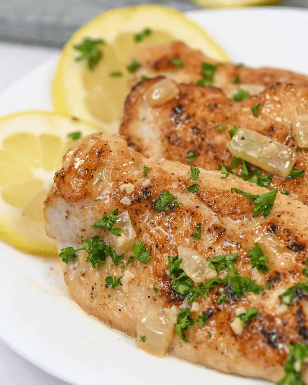 Lemon Garlic Chicken: Grilled chicken breasts topped with a zesty lemon-garlic sauce and garnished with fresh parsley, complemented by lemon slices in the background on a pristine white plate.
