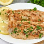 A plate with three pieces of seasoned grilled fish garnished with chopped parsley and onions, accompanied by thin lemon slices reminiscent of our Lemon Garlic Chicken in the background.