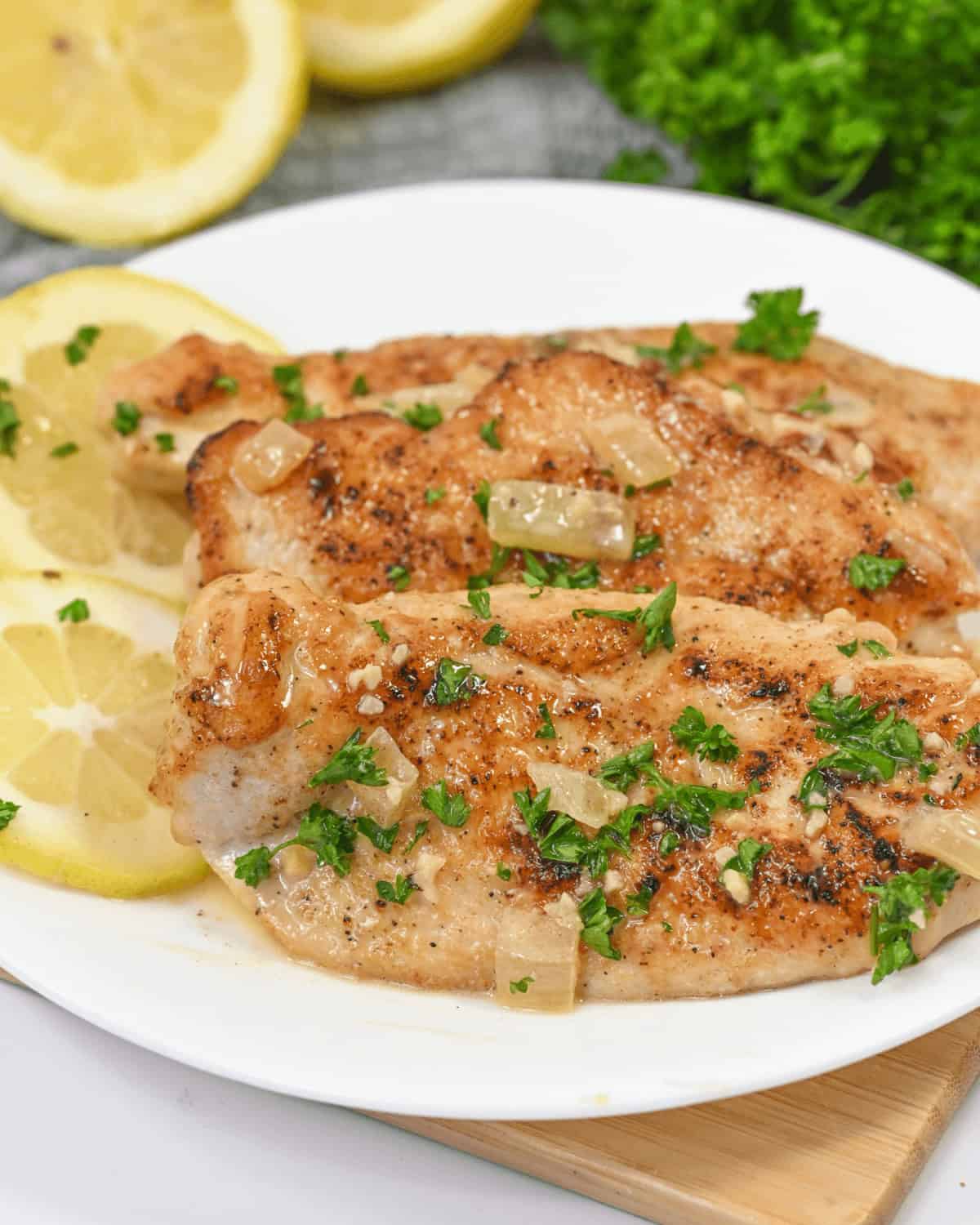 A plate with three pieces of seasoned grilled fish garnished with chopped parsley and onions, accompanied by thin lemon slices reminiscent of our Lemon Garlic Chicken in the background.