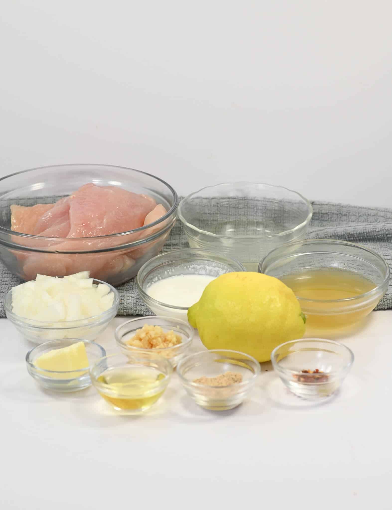 An assortment of cooking ingredients for Lemon Garlic Chicken arranged on a counter, including raw chicken, chopped onions, lemon, garlic, oil, butter, lemon juice, spices, and various liquids in small bowls.