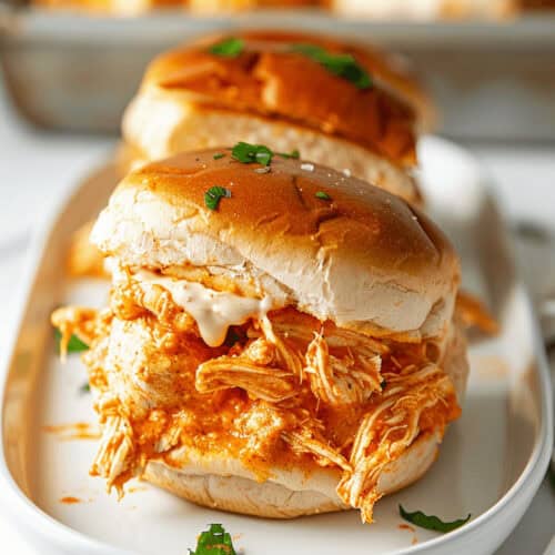 Buffalo chicken sliders topped with a creamy sauce on a bun, elegantly garnished with parsley, and served on a white plate.