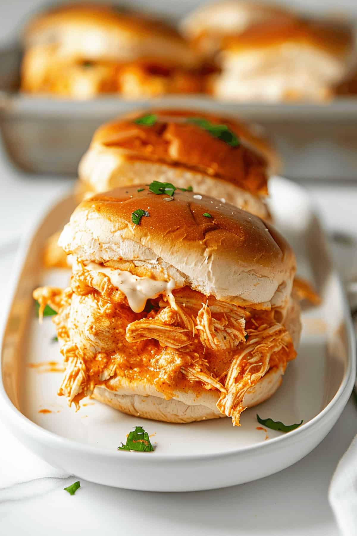 Buffalo chicken sliders topped with a creamy sauce on a bun, elegantly garnished with parsley, and served on a white plate.