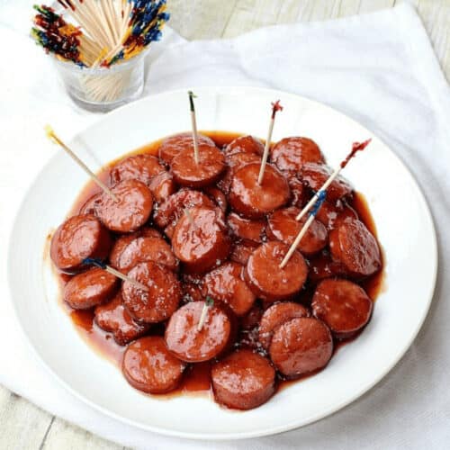A plate of slow cooker glazed cocktail sausages with toothpicks, next to a glass of colorful party picks on a wooden table.