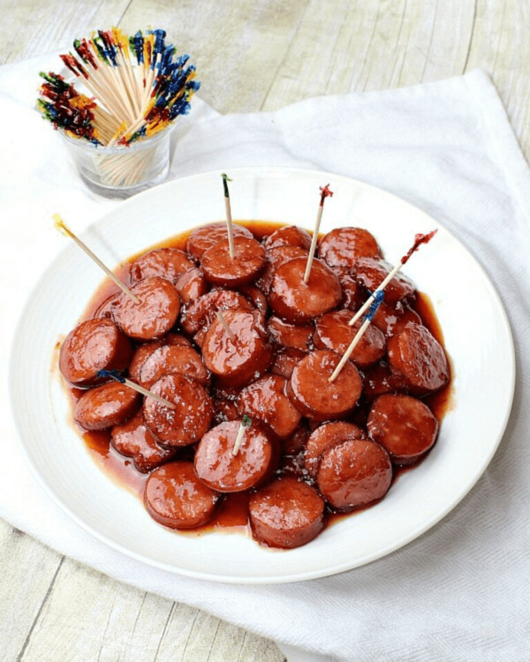 A plate of slow cooker glazed cocktail sausages with toothpicks, next to a glass of colorful party picks on a wooden table.