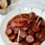 A hand holding a cocktail stick with a glazed meatball over a plate of slow cooker cocktail sausages in a shiny sauce.