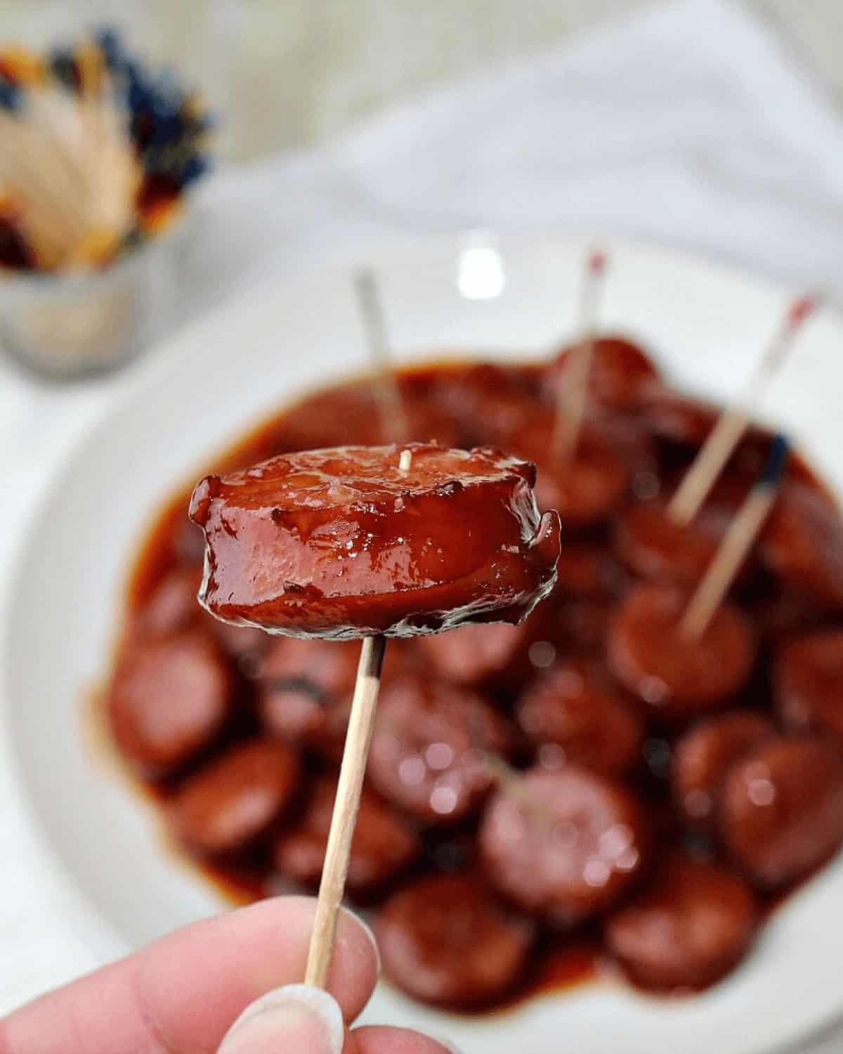 A hand holding a cocktail stick with a glazed meatball over a plate of slow cooker cocktail sausages in a shiny sauce.
