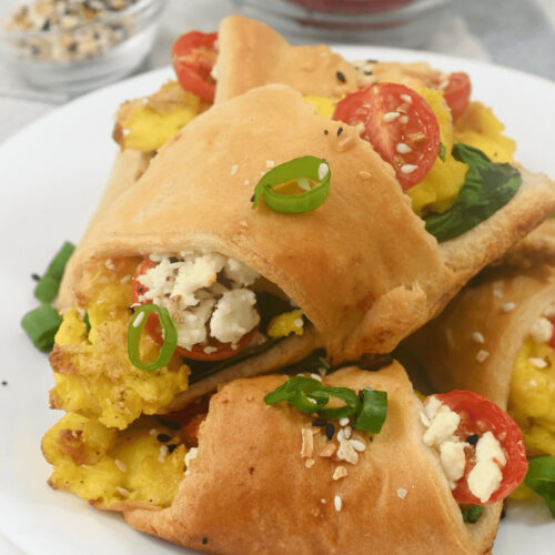 A plate of breakfast croissant roll ups stuffed with feta, cherry tomatoes, and green onions, garnished with seeds.