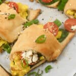 A breakfast croissant stuffed with scrambled eggs, spinach, cherry tomatoes, and crumbled feta, garnished with scallions and sesame seeds.