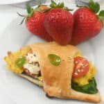 A piece of the dish on a white plate with fresh strawberries. tomatoes, greens, and eggs on a white plate, garnished with strawberries and cereal in the background.