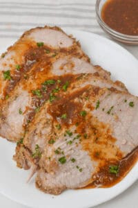 Slices of roasted crock pot pork tenderloin topped with sauce and garnished with herbs on a white plate. Nearby is a small bowl containing additional sauce.