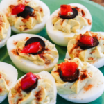 A plate of deviled eggs without mayo, garnished with paprika and a slice of olive and red pepper.