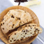A loaf on a wicker plate with one slice cut. Nearby, a measuring cup with milk, butter, and raisins sits on a gray cloth—a comforting scene reminiscent of making easy Irish soda bread.