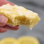 Close-up of a hand holding a bitten dessert revealing a moist and crumbly texture. Two lemon slices are blurred in the background.
