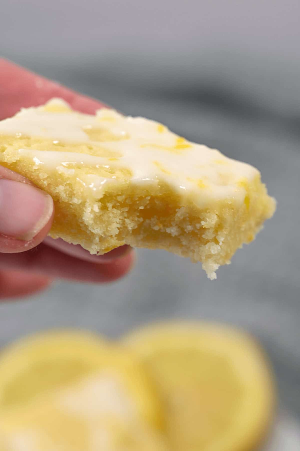 Close-up of a hand holding a bitten dessert revealing a moist and crumbly texture. Two lemon slices are blurred in the background.
