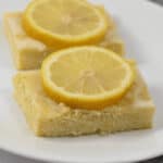 Two lemon bars, reminiscent of delightful lemon brownies, rest on a white plate, each topped with a glistening slice of lemon.
