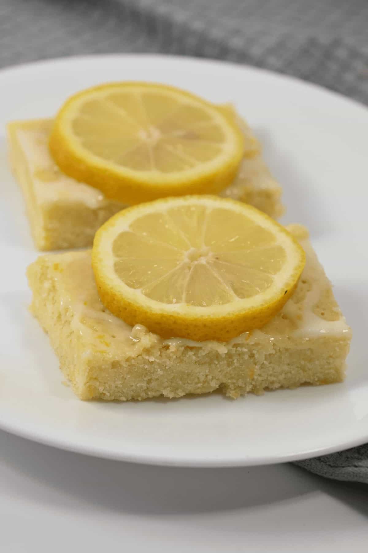 Two lemon bars, reminiscent of delightful lemon brownies, rest on a white plate, each topped with a glistening slice of lemon.