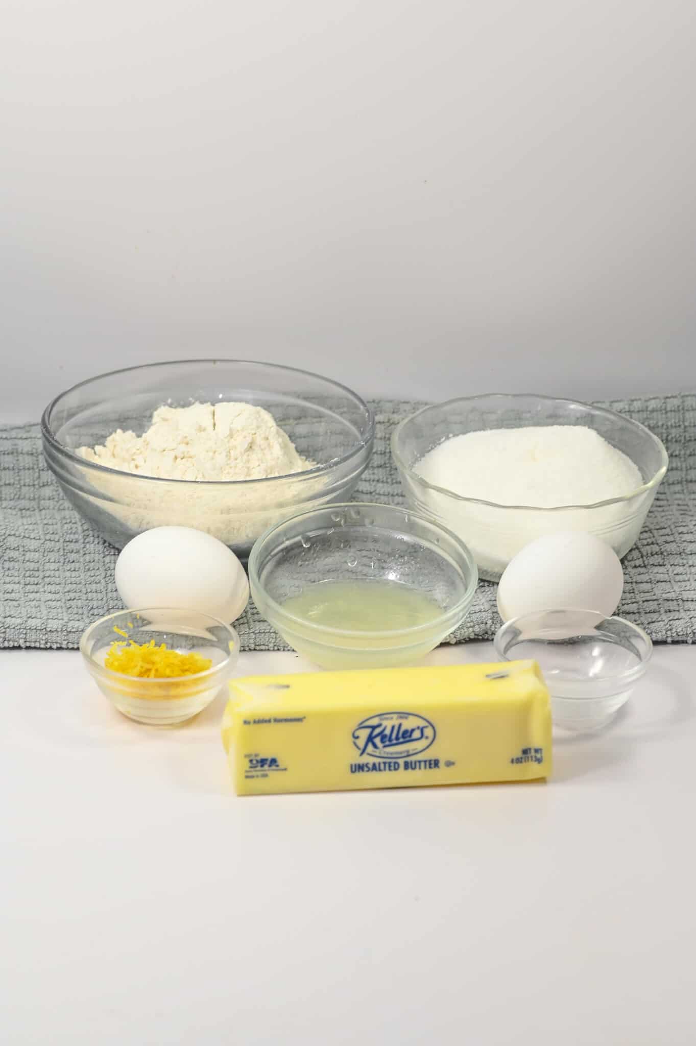 Baking ingredients for the bars are beautifully arranged on a table: flour, sugar, eggs, lemon juice, lemon zest, vanilla extract, and a stick of unsalted butter.