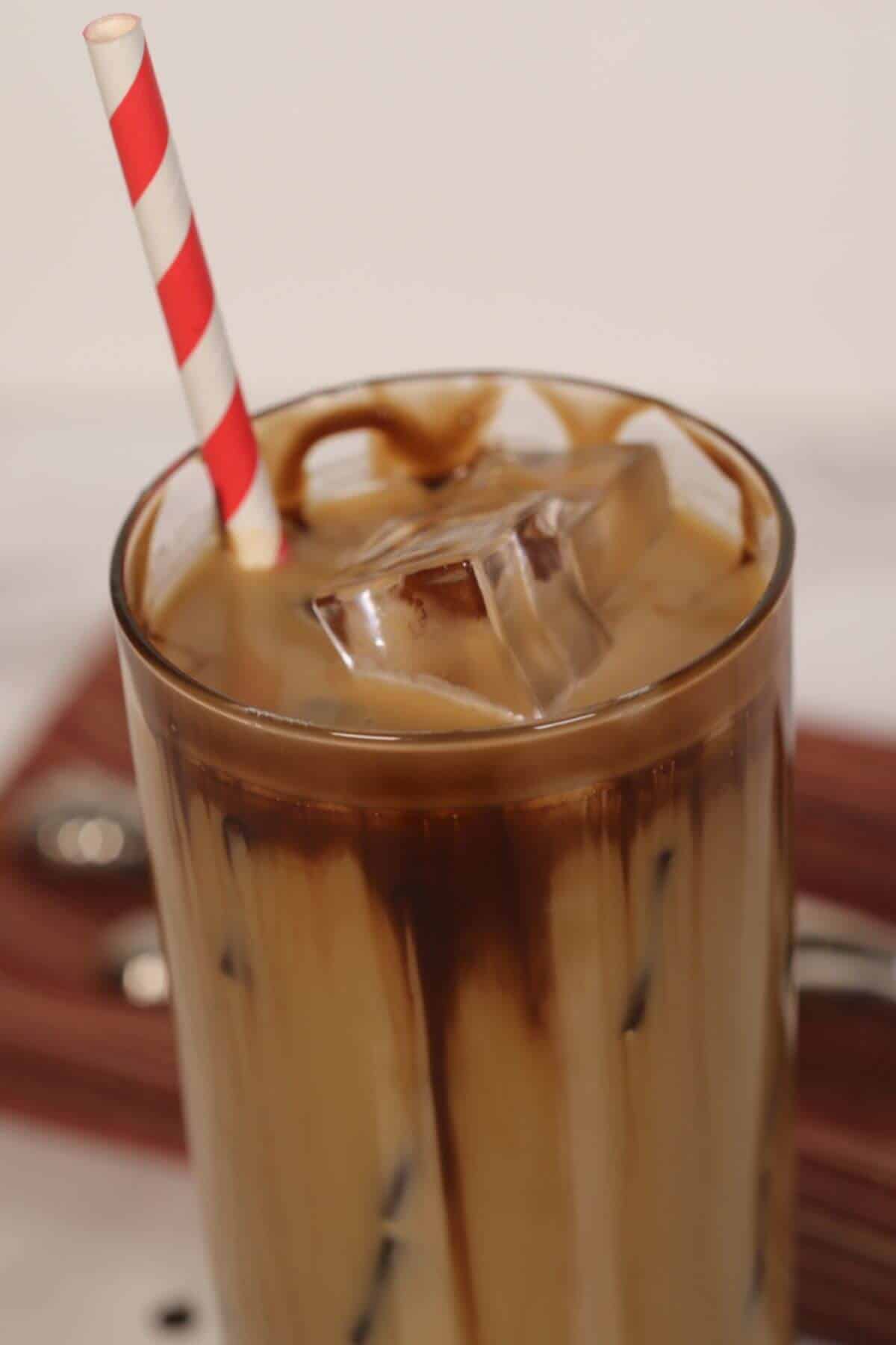 A glass of caramel iced coffee with milk and chocolate drizzle, filled with ice cubes, and topped off with a red-and-white striped straw.