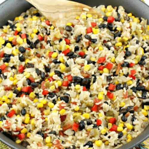 A skillet filled with a mixture of cooked rice, black beans, corn, diced red peppers, and onions—a perfect ensemble for Mexican Rice and Beans—with a wooden spoon resting on the side.