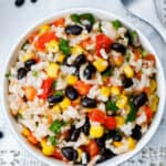 A bowl of Mexican Rice and Beans salad with black beans, corn, tomatoes, and cilantro sits on a white lace-patterned tablecloth with a fork beside it.