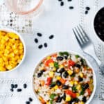 A bowl of Mexican Rice and Beans mixed with corn, black beans, tomatoes, and herbs sits alongside a fork. Surrounding dishes contain additional black beans, corn, and a glass of water. Black beans are scattered on the tablecloth.