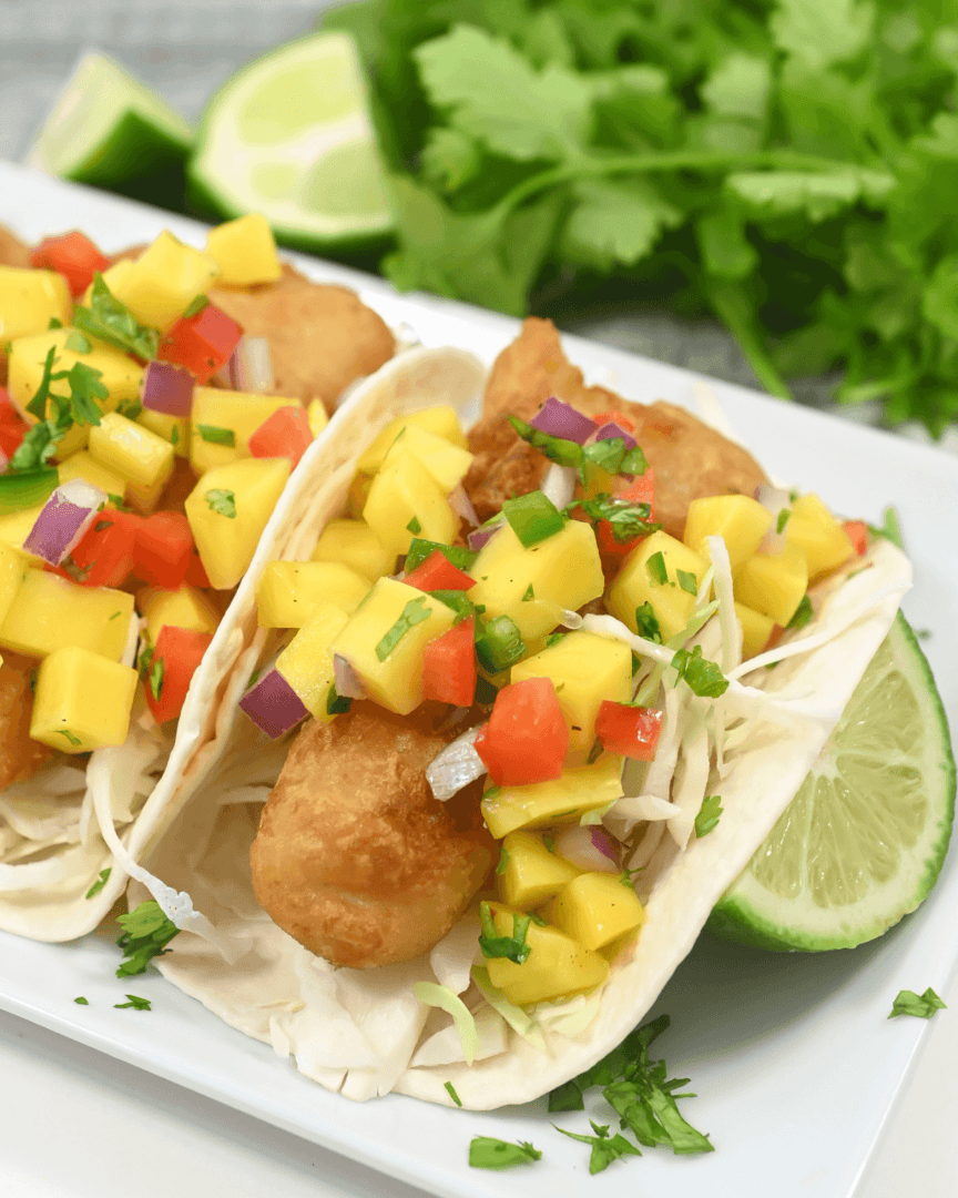 Fish tacos with mango salsa, cabbage, and lime on a white plate garnished with chipotle chicken salad.