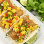 Fish tacos with mango salsa, garnished with cilantro and lime wedges on a white plate.