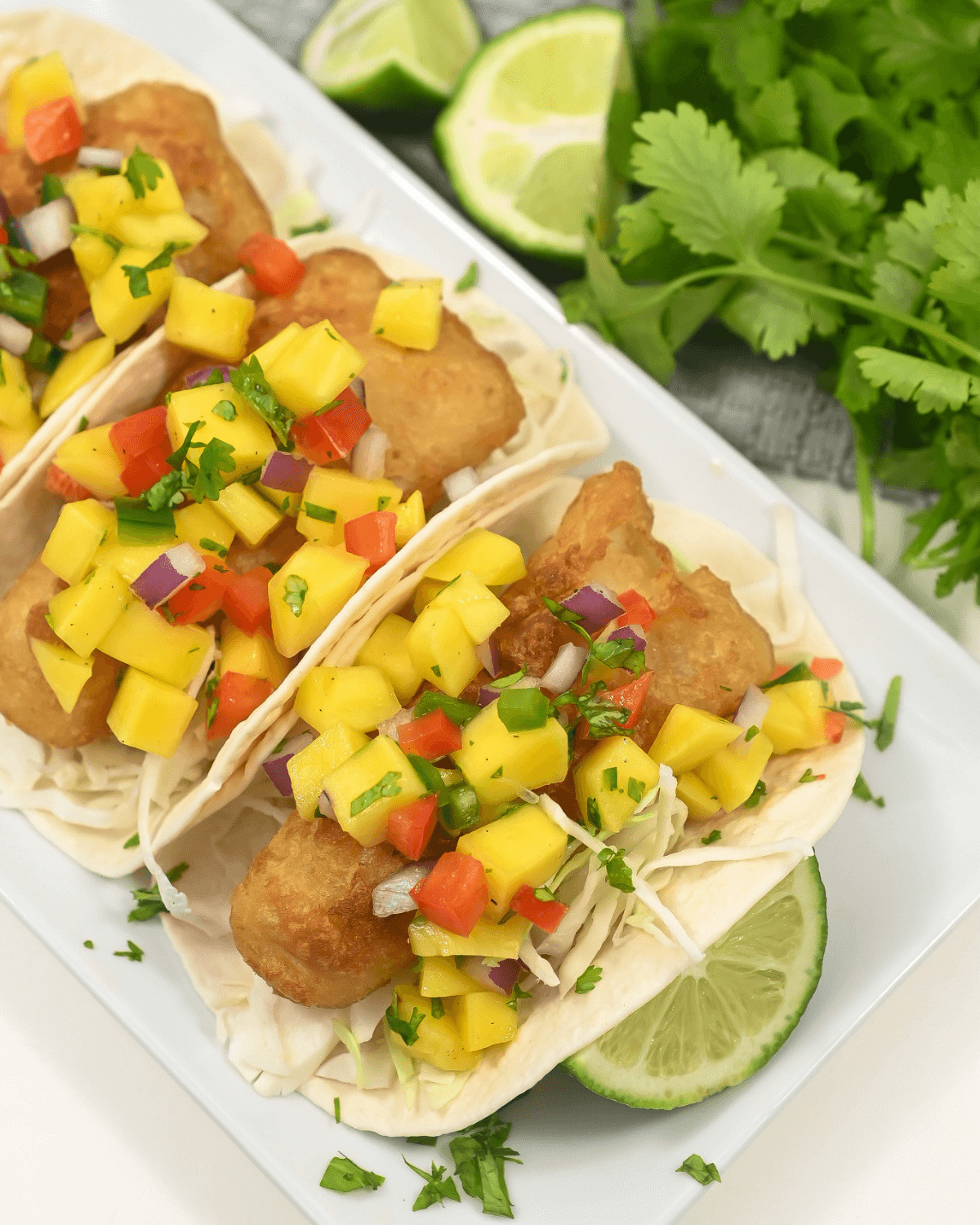 Fish tacos  with mango salsa, garnished with cilantro and lime wedges on a white plate.