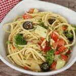 A bowl of spaghetti pasta salad with broccoli, diced tomatoes, olives, and herbs on a wooden surface, with a red and white cloth napkin beside it.