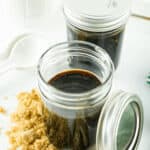 Two jars of homemade honey teriyaki sauce, one open and one closed, on a white surface with brown sugar and ginger scattered around.
