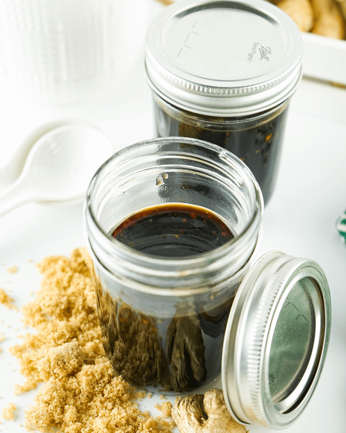 Two jars of homemade honey teriyaki sauce, one open and one closed, on a white surface with brown sugar and ginger scattered around.