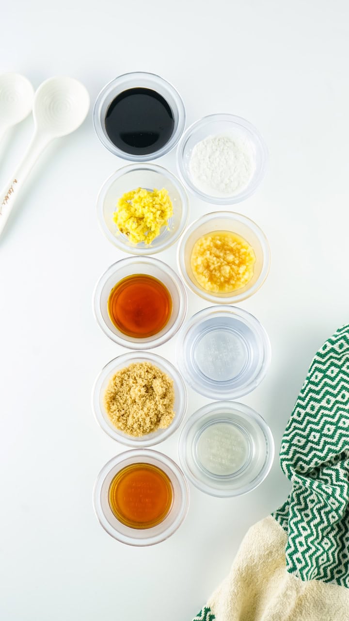 Assorted ingredients in small clear containers on a white surface, with measuring spoons to the left and a green towel to the right, including honey teriyaki sauce.