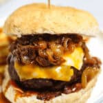 A close-up of a Whiskey Burger with melted cheese, caramelized onions, and a beef patty in a bun, held together by a toothpick, on a white plate.
