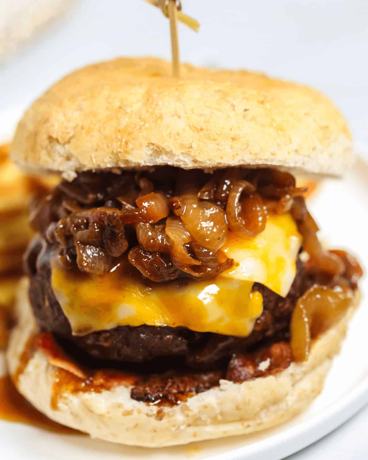 A close-up of a Whiskey Burger with melted cheese, caramelized onions, and a beef patty in a bun, held together by a toothpick, on a white plate.
