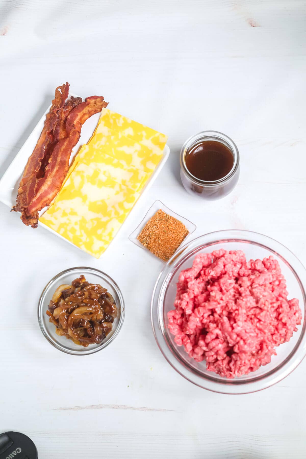 A variety of Whiskey Burger ingredients displayed on a white surface: cooked bacon, cheese slices, a bowl of ground beef, a glass jar of sauce, a bowl of cooked onions, and a small container of seasoning.