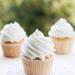 Three vanilla cupcakes with white swirled frosting sit on a light surface; one cupcake, topped with vanilla buttercream frosting from our favorite recipe, is placed on a white doily in the foreground.