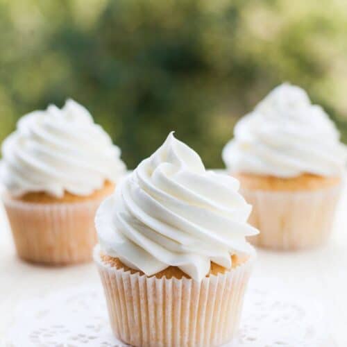 Three vanilla cupcakes with white swirled frosting sit on a light surface; one cupcake, topped with vanilla buttercream frosting from our favorite recipe, is placed on a white doily in the foreground.