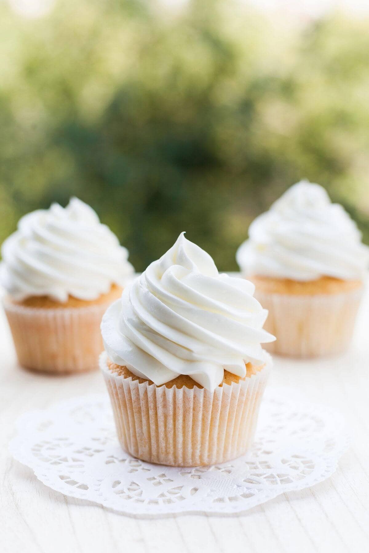 Three vanilla cupcakes with white swirled frosting sit on a light surface; one cupcake, topped with vanilla buttercream frosting from our favorite recipe, is placed on a white doily in the foreground.