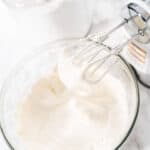 A glass bowl filled with icing sits on a marble surface next to a jar of sugar and an electric hand mixer with beaters covered in whipped cream, perfect for starting your favorite dessert.