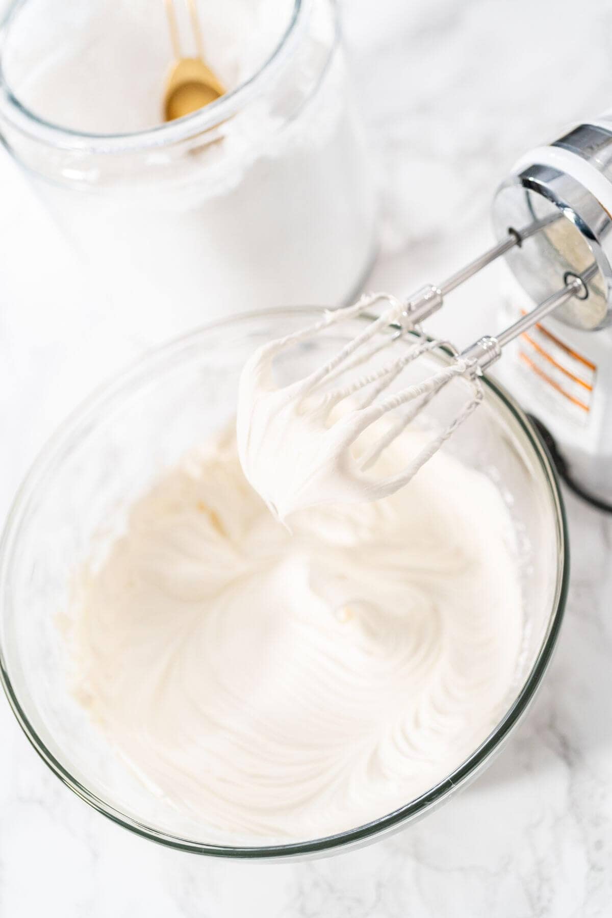 A glass bowl filled with icing sits on a marble surface next to a jar of sugar and an electric hand mixer with beaters covered in whipped cream, perfect for starting your favorite dessert.