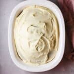 Oval white dish filled with a cake topped with smooth, swirled vanilla buttercream frosting, set on a light surface next to a pink cloth—perfect inspiration for your next Vanilla Buttercream Frosting Recipe.