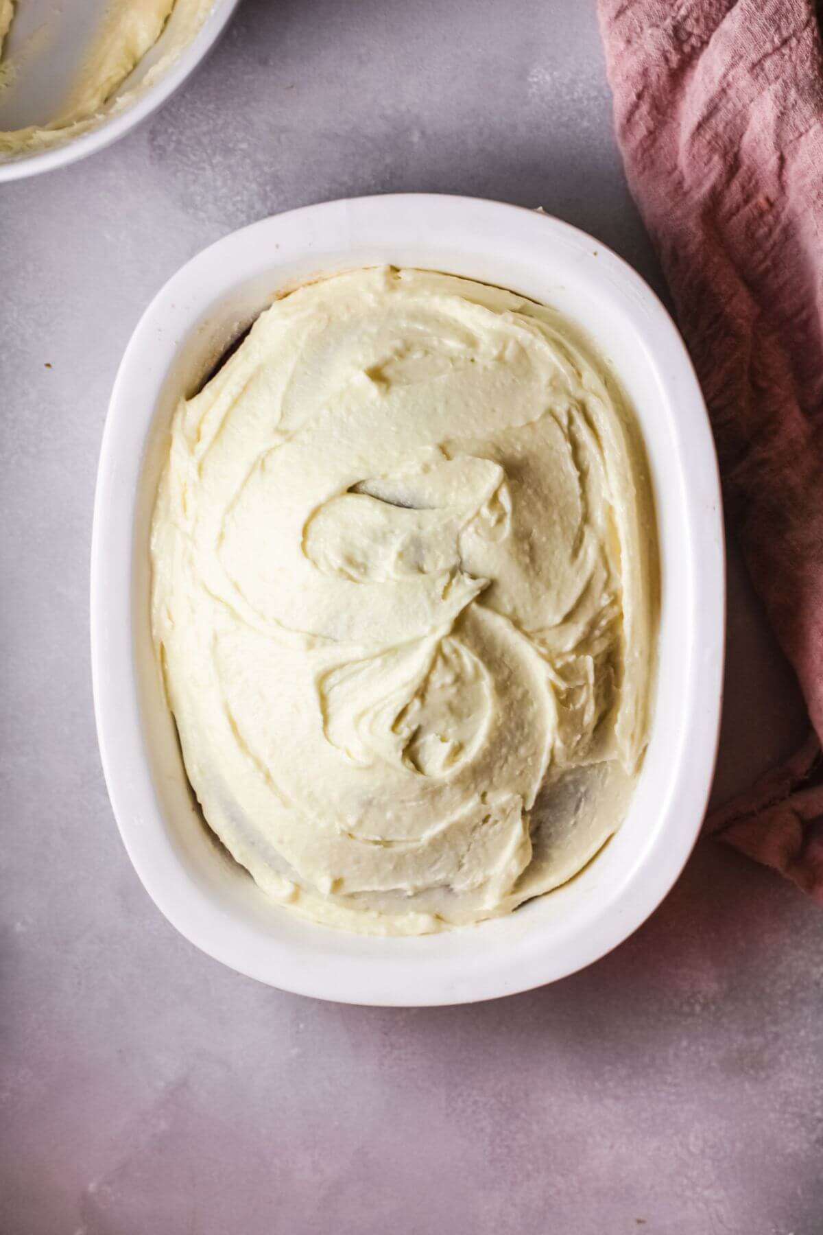 Oval white dish filled with a cake topped with smooth, swirled vanilla buttercream frosting, set on a light surface next to a pink cloth—perfect inspiration for your next Vanilla Buttercream Frosting Recipe.