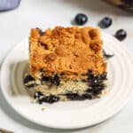 A slice of blueberry crumb cake sits on a white plate next to a fork. The cake has a crumbly topping and visible blueberries. A baking dish with more cake is in the background.