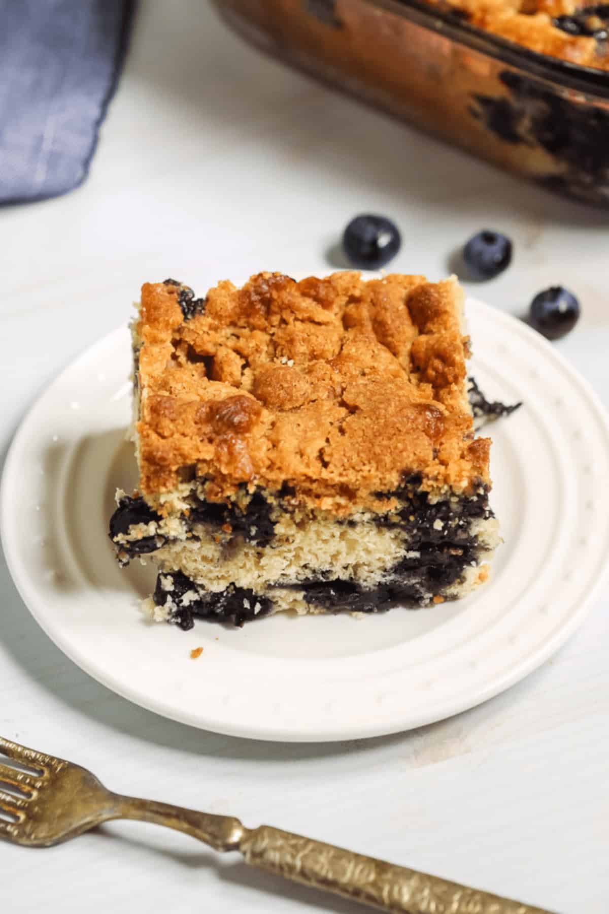 A slice of blueberry crumb cake sits on a white plate next to a fork. The cake has a crumbly topping and visible blueberries. A baking dish with more cake is in the background.