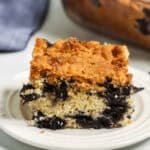 A slice of blueberry crumb cake on a white plate with a glass baking dish containing more cake in the background.