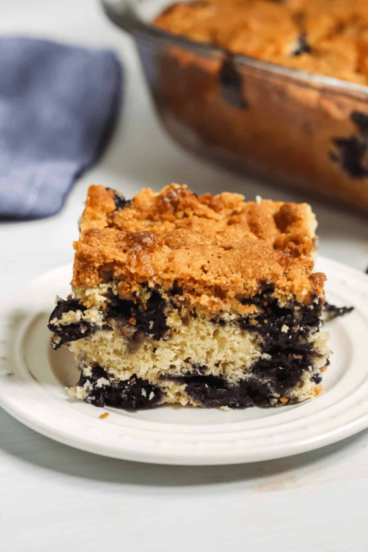 A slice of blueberry crumb cake on a white plate with a glass baking dish containing more cake in the background.
