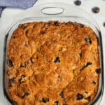 A square glass baking dish holding a freshly baked Blueberry Crumb Cake rests on a light surface with scattered blueberries and a folded blue cloth nearby.