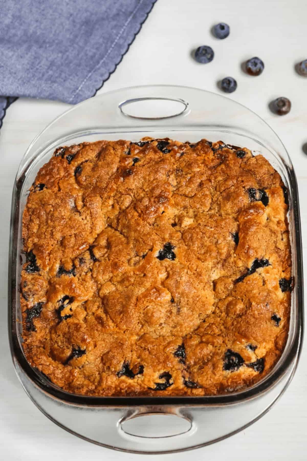 A square glass baking dish holding a freshly baked Blueberry Crumb Cake rests on a light surface with scattered blueberries and a folded blue cloth nearby.