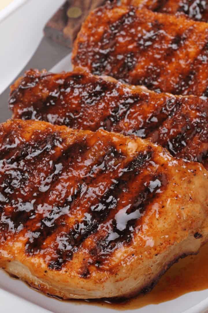 Grilled pork chops, marinated to perfection, boast a glossy, caramelized coating as they are arranged in a row on a white plate.